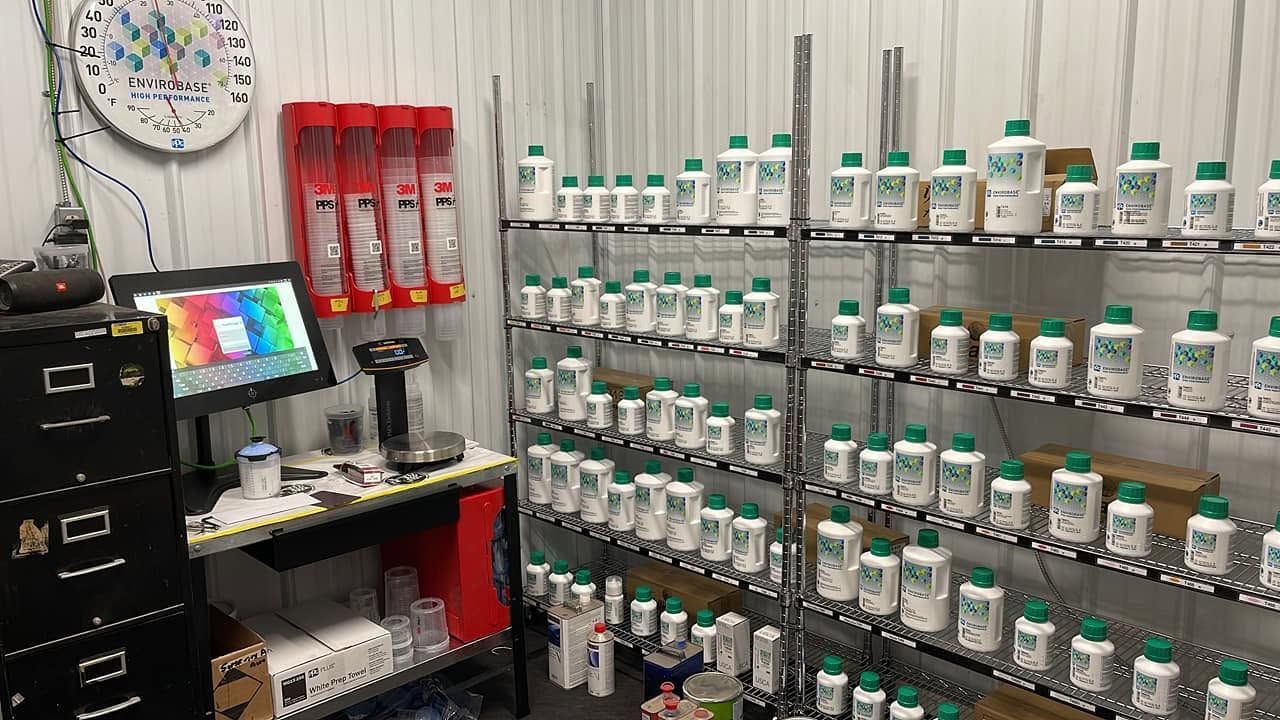 Laboratory shelf with rows of labeled chemical bottles, a laptop, and storage racks in a small workspace