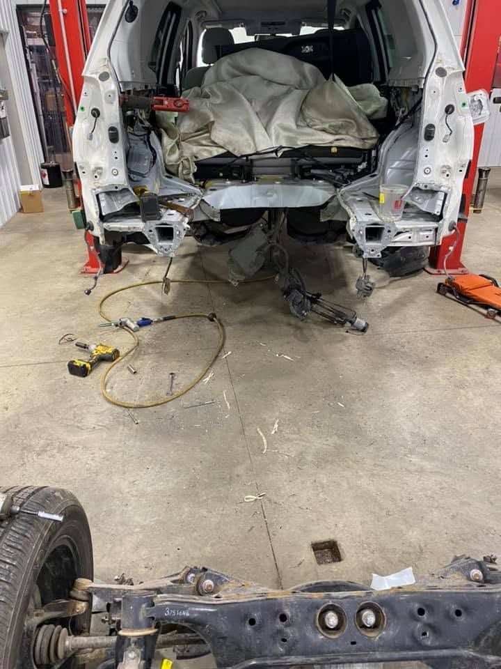 Vehicle in a repair shop with rear interior stripped and detached front subframe visible on the floor.