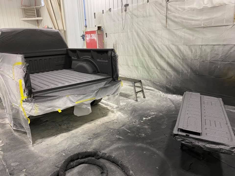 Pickup truck in a paint booth, masked with plastic and paper, with detached parts on the floor.