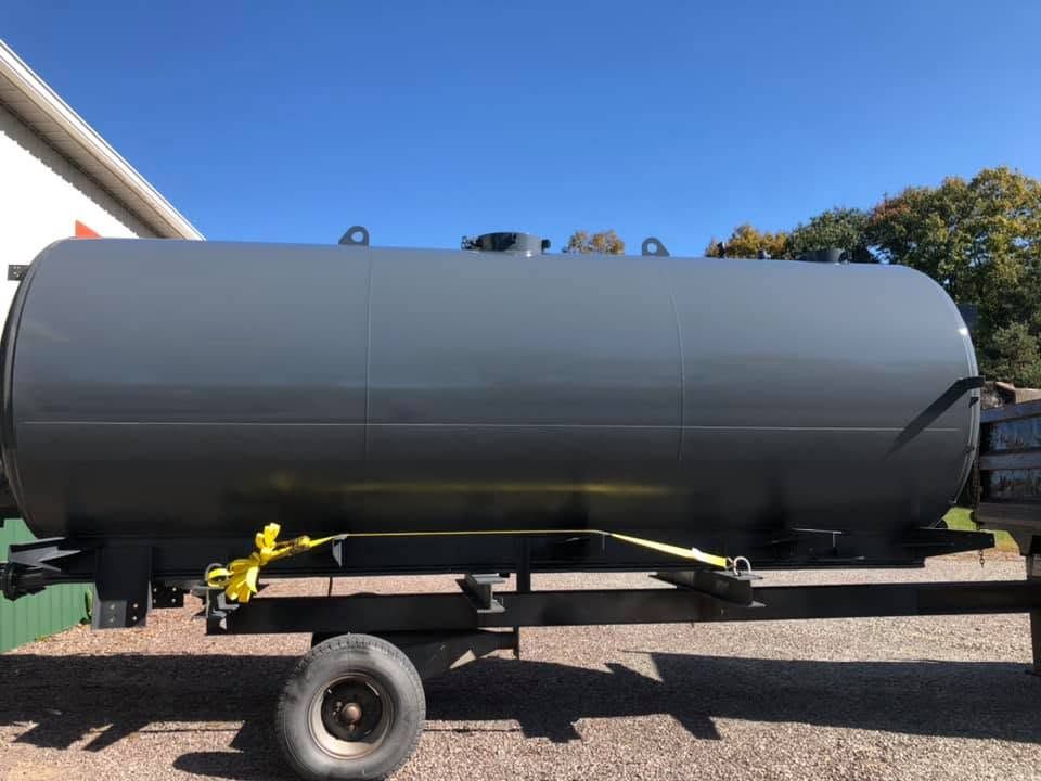 Large gray tanker trailer on a flatbed under a blue sky, with yellow straps and a single wheel visible.