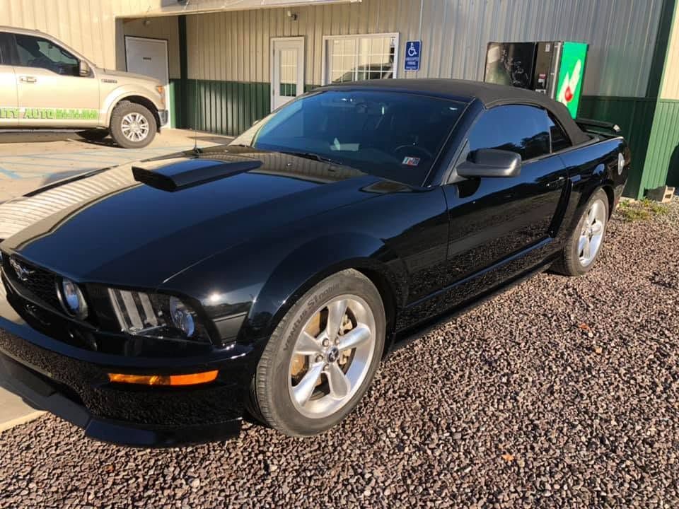 Black Ford Mustang convertible parked on gravel outside a building