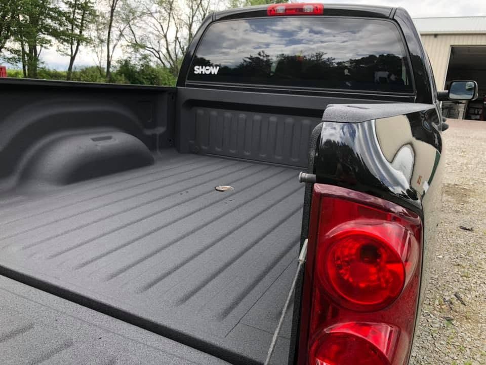 Black pickup truck bed and rear taillight, viewed from the tailgate in an outdoor gravel area.