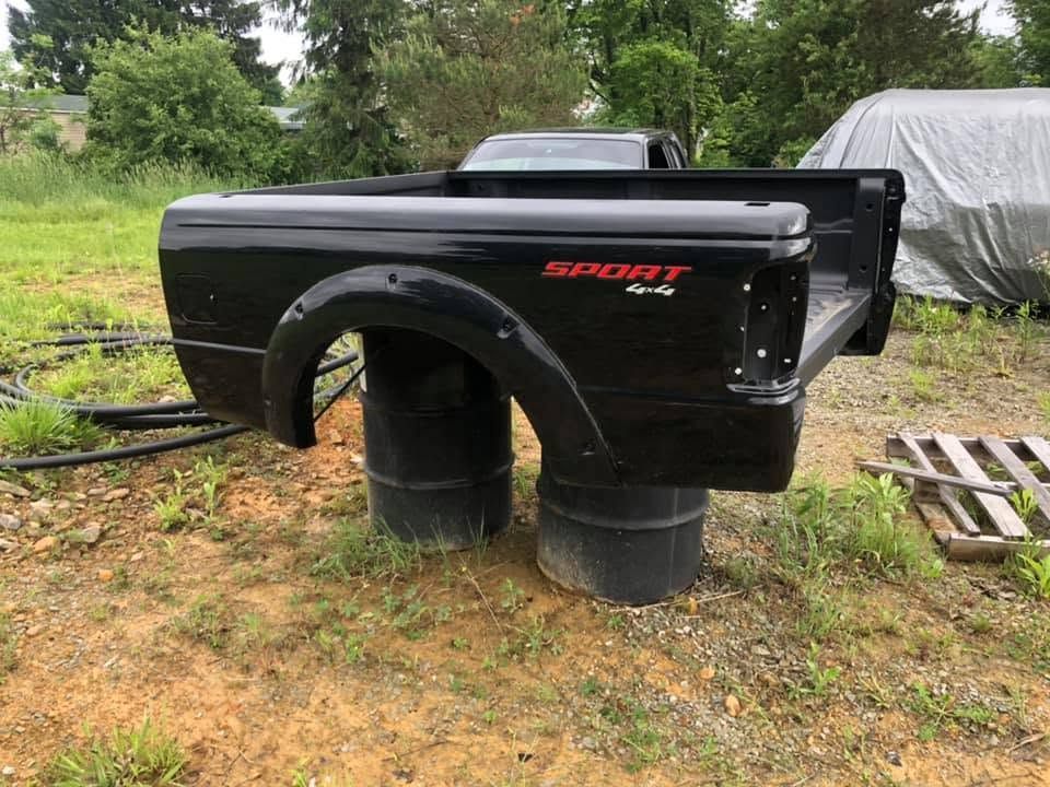 Black truck bed/tub shell on two barrels outdoors, with red decal and yard equipment nearby.