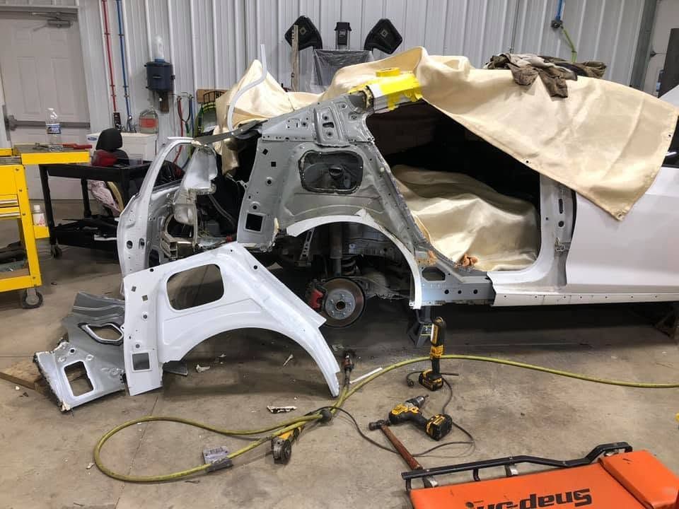 Damaged white car body in an auto repair shop, with detached panels and tools on the floor.