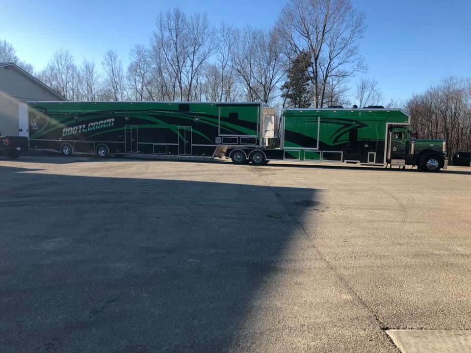 Two green semi-trucks parked in a lot beside a building, under bare trees on a clear day