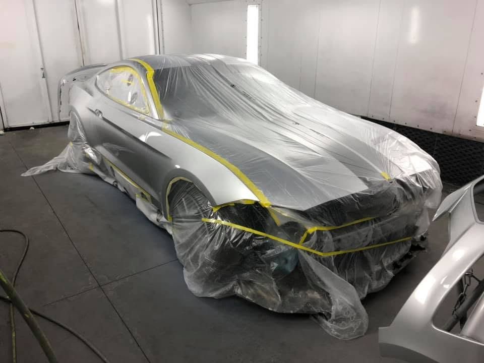Car covered in plastic masking inside a spray booth, with yellow tape outlining panels.