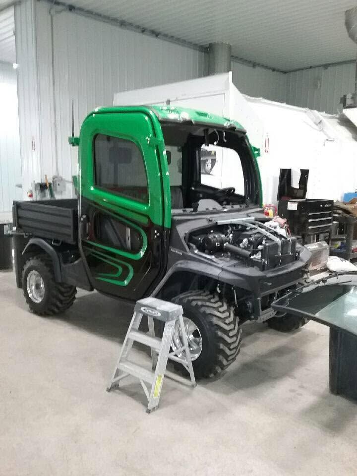 Green off-road utility vehicle in a garage, front panel removed during repair, with a step stool nearby