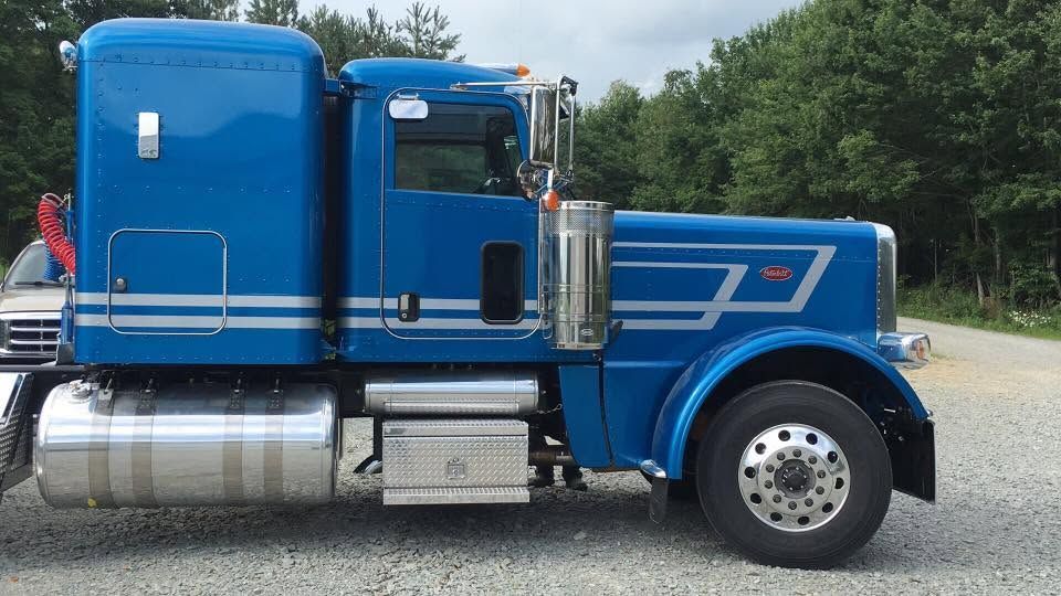 Blue semi-truck cab parked on gravel, side view with chrome fuel tanks and white racing stripes.