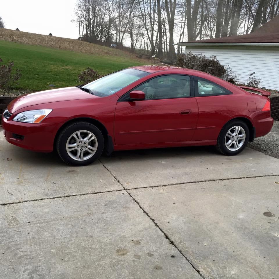 Red coupe parked on a driveway beside a white garage, with a grassy hill in the background.