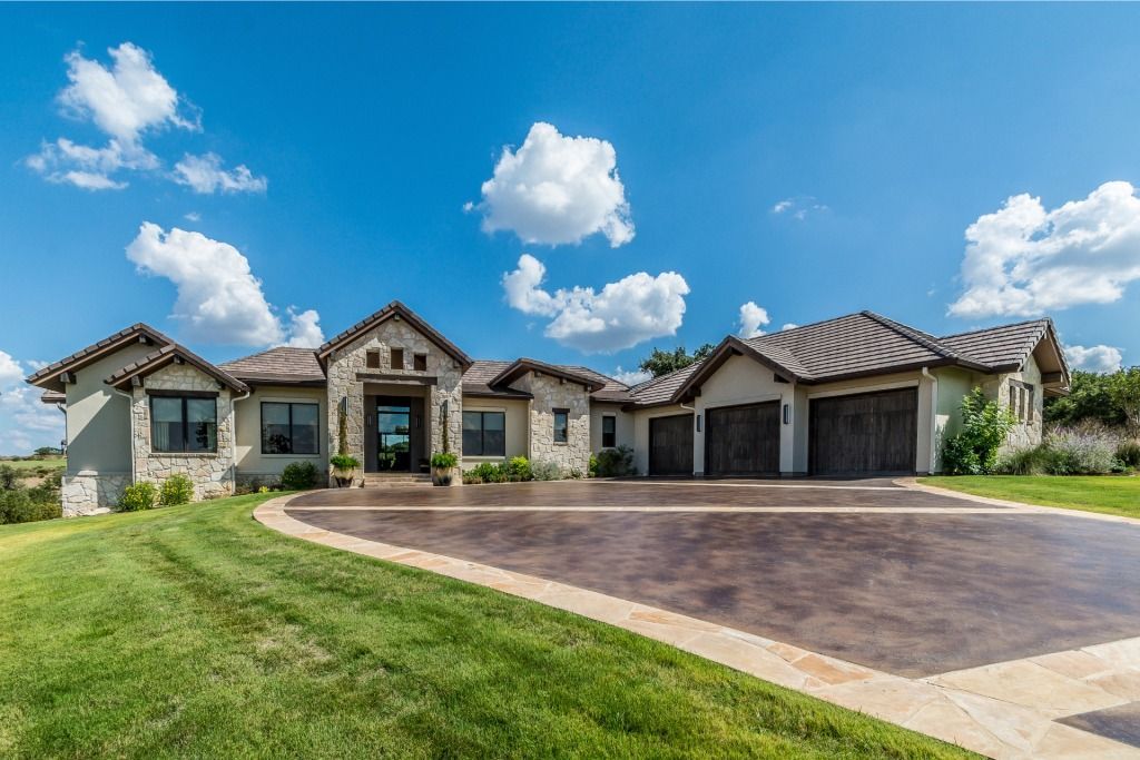 Sprawling beige house with curved driveway and three-car garage under a bright blue sky with fluffy clouds.