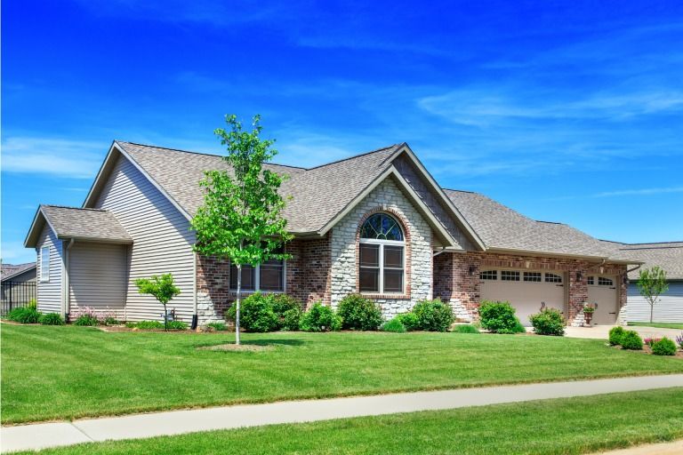House with beige siding, brick facade, arched window, and attached garage under a bright blue sky.