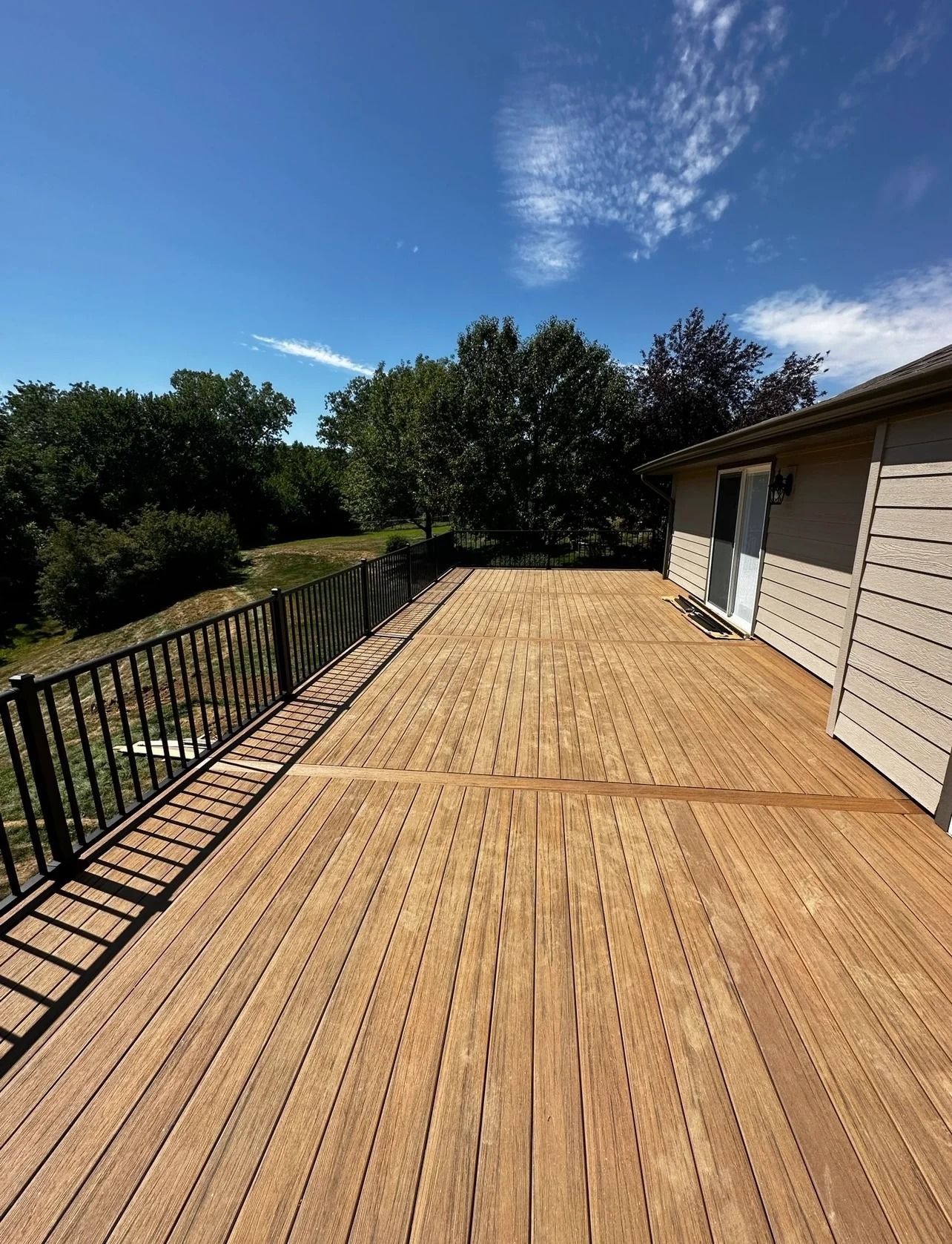 Wooden deck with black railing next to house; sunny day with blue sky and trees.