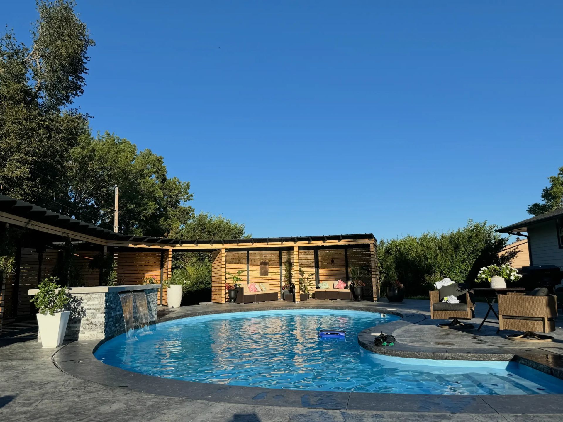 A backyard pool scene with wooden cabana, seating, and a clear blue sky.