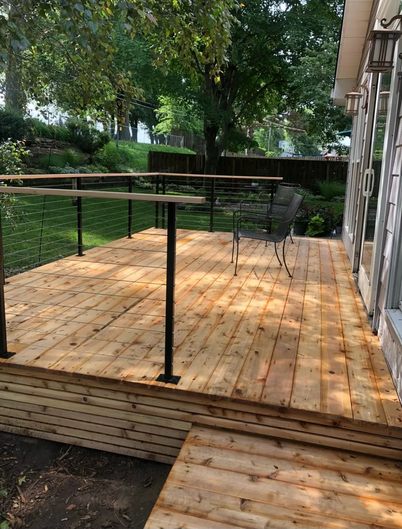 Wooden deck with black railings and cable wires, a chair, and a ramp, outdoors with trees.