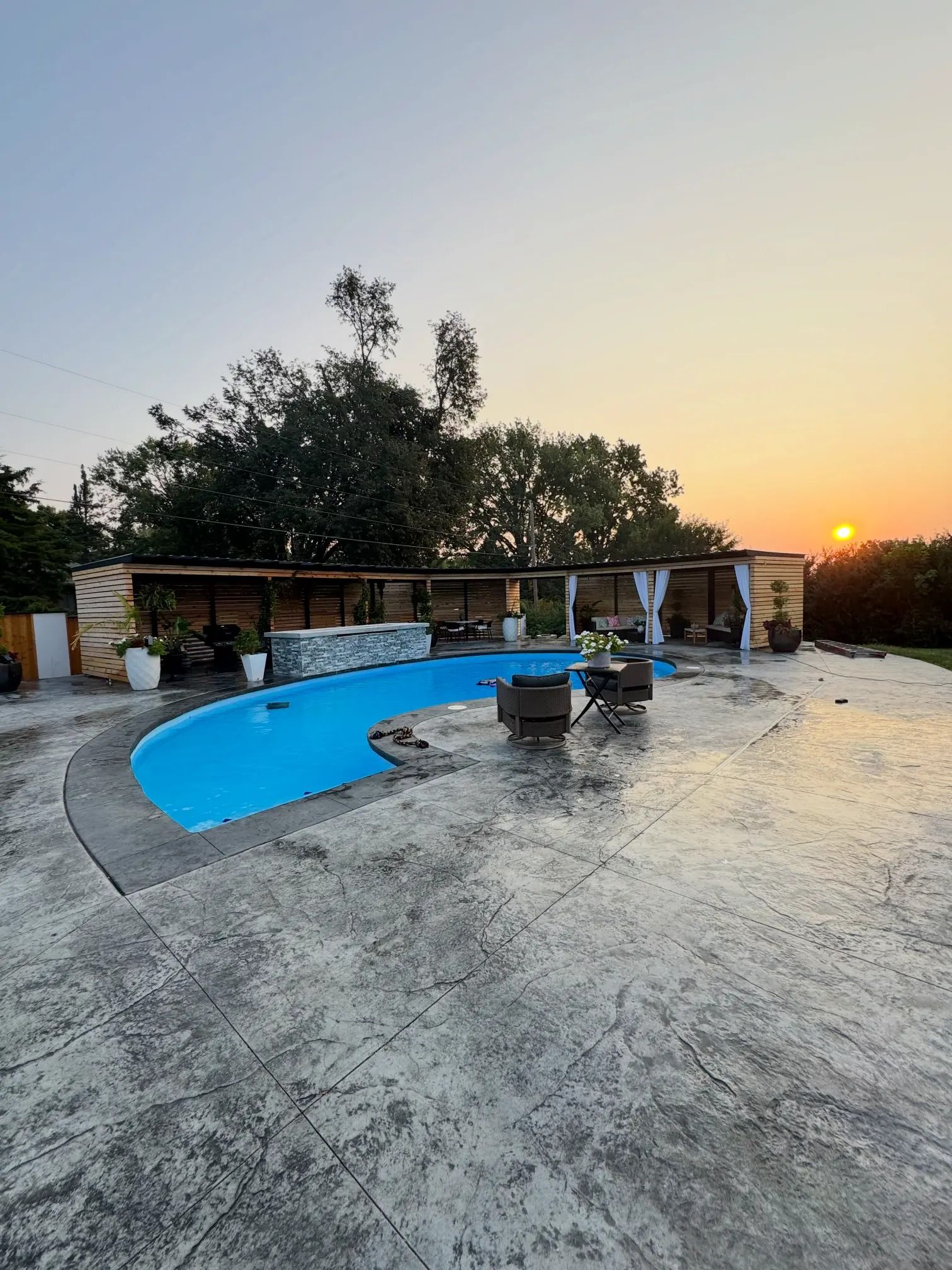 Pool with blue water and stone patio at sunset. Stone gazebo with white curtains.