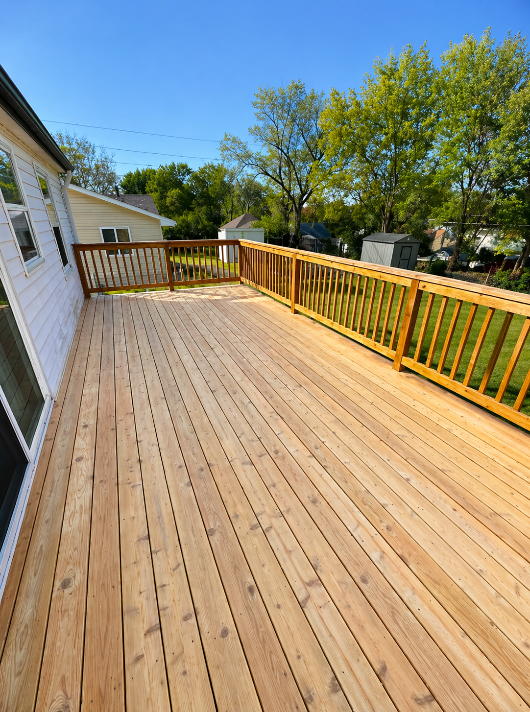 Wide wooden backyard deck with yellow railings beside a white house under a blue sky