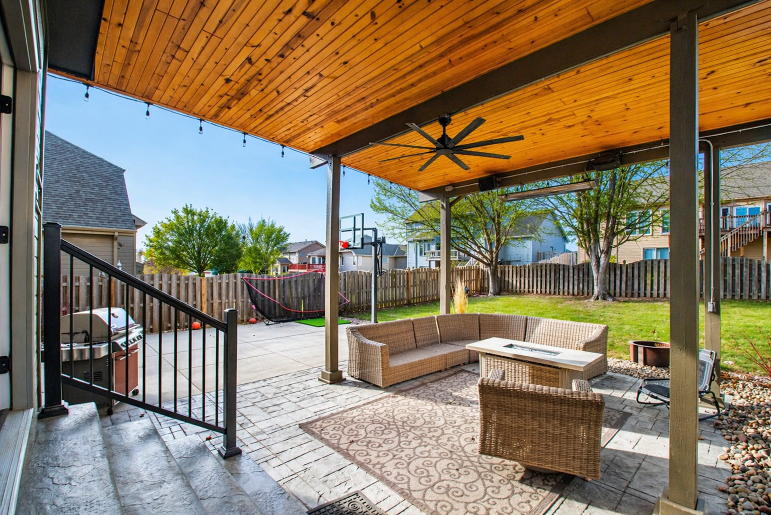 Covered backyard patio with wicker seating, ceiling fan, and stone flooring overlooking a fenced yard