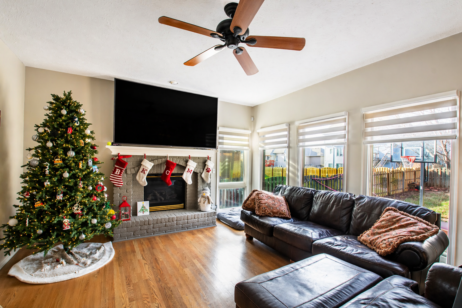 Christmas living room with decorated tree, fireplace, leather sofa, and large windows overlooking a backyard