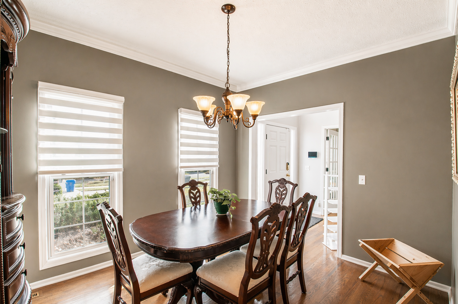 Bright dining room with round wooden table, six chairs, beige walls, and two windows with blinds.