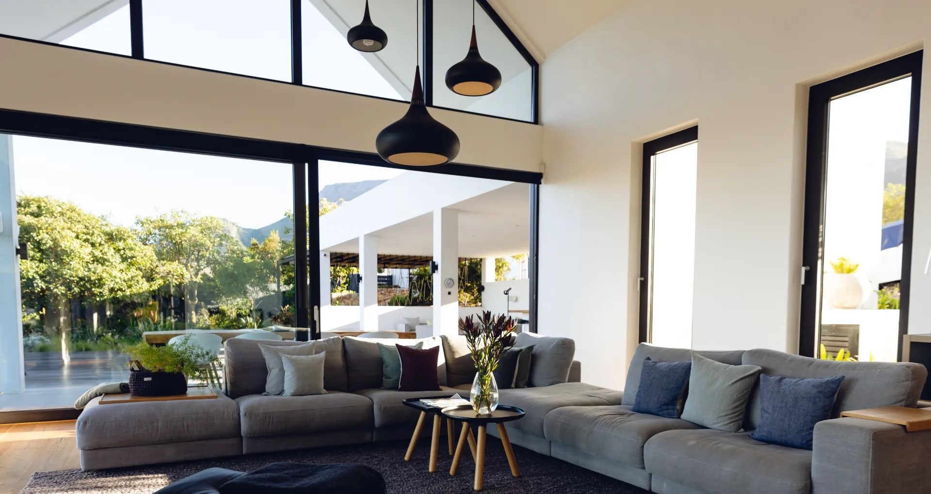 Spacious modern living room with gray sectional sofa, large windows, and pendant lights overlooking outdoor view.