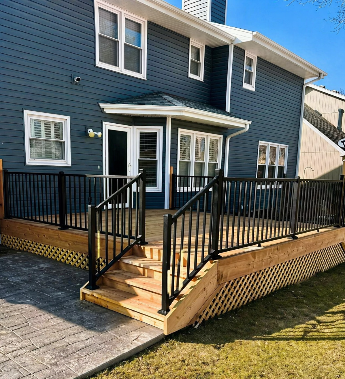 Blue house with white trim, front deck, black railings, and brick-paved driveway under a clear sky
