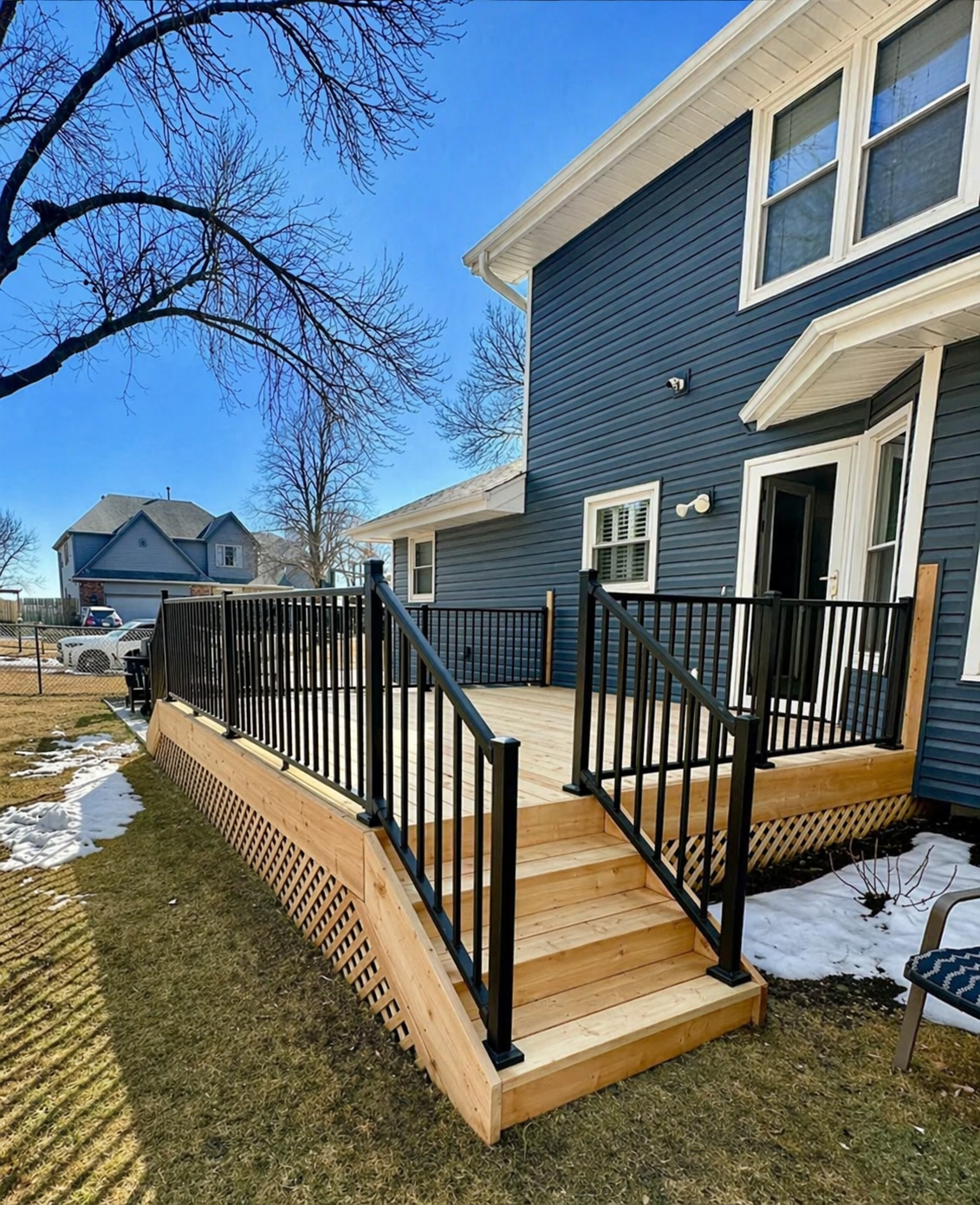 Raised wooden deck stairs with black railings attached to a blue house in a snowy yard