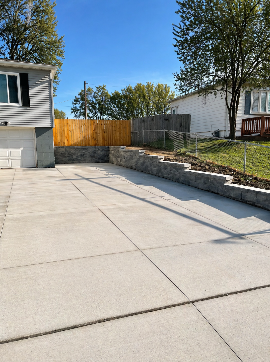 Concrete driveway beside a house, stone retaining wall, wooden fence, and grassy yard under blue sky