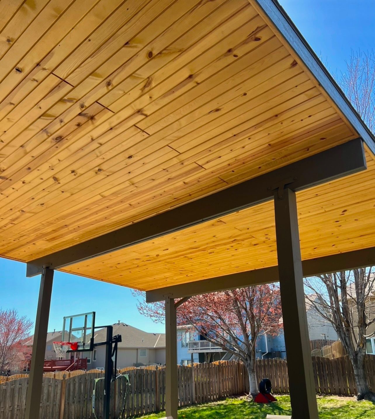 Covered backyard patio with wood plank ceiling, dark posts, and a basketball hoop nearby.