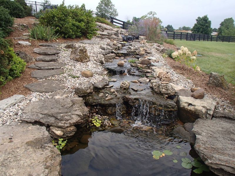 Stone waterfall flowing into a pond, with stepping stones, landscaping, and a grassy field.
