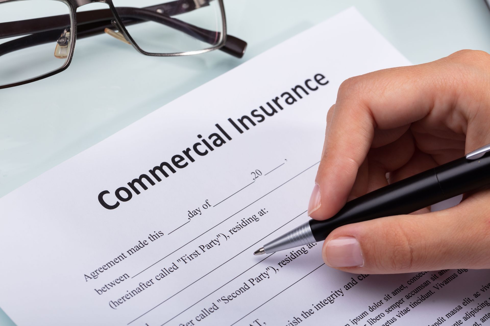 Hand signing a commercial insurance form beside eyeglasses on a desk