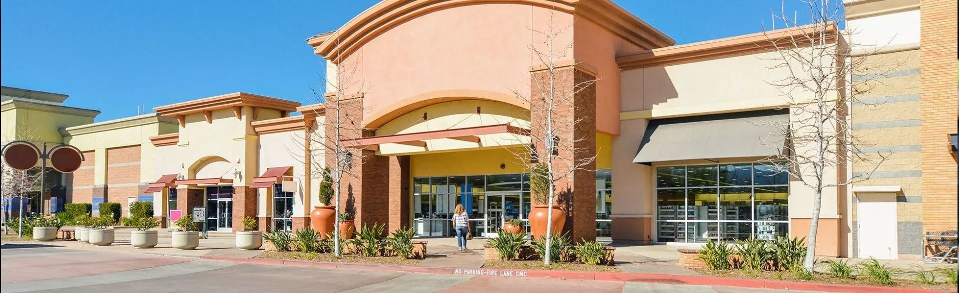 Outdoor shopping center with storefronts, tan stone фасades, and a central peach-colored archway under a blue sky