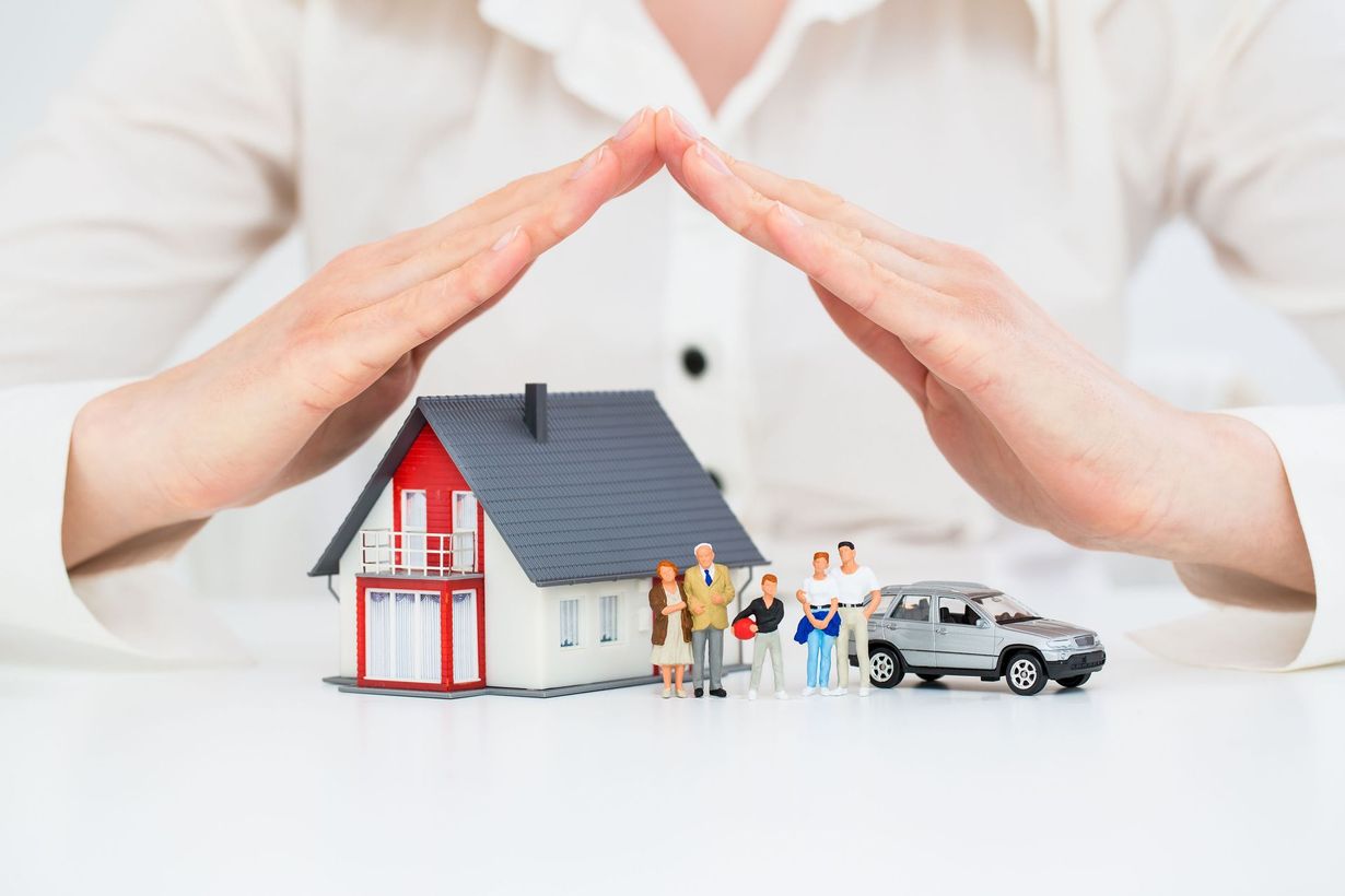 Hands protecting a house, family figurines, and a car on a white surface