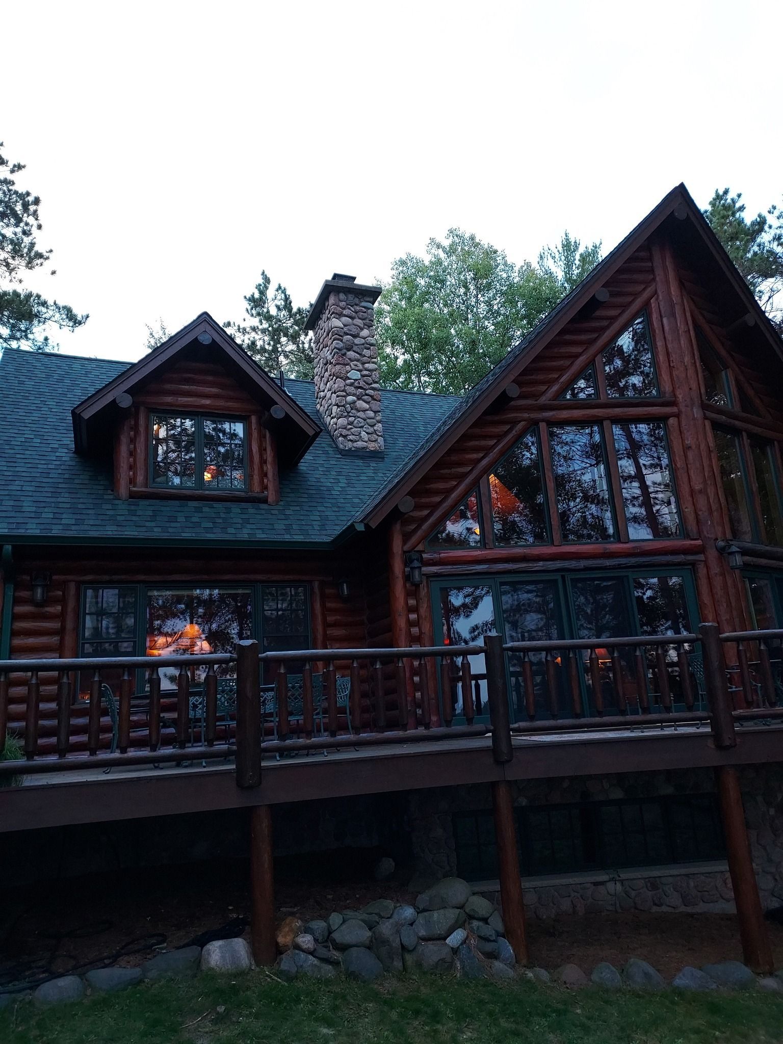 Log cabin with a wooden deck, stone chimney, and large windows reflecting interior lights.