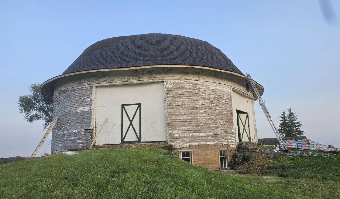 Round barn with a dark roof, white walls, and green field. Ladders are present.