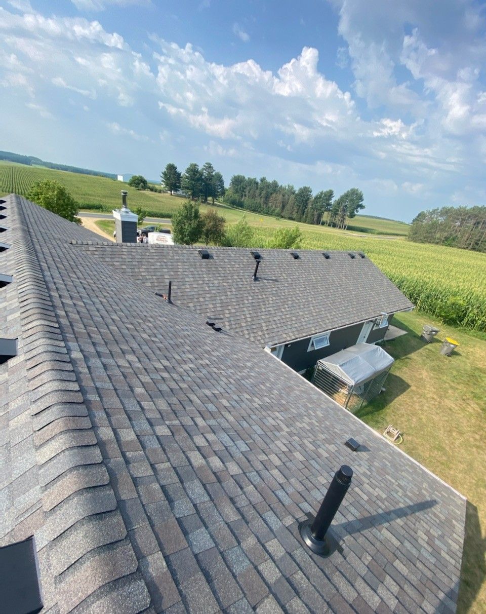 View of a grey-shingled roof with a field and sky in the background on a sunny day.