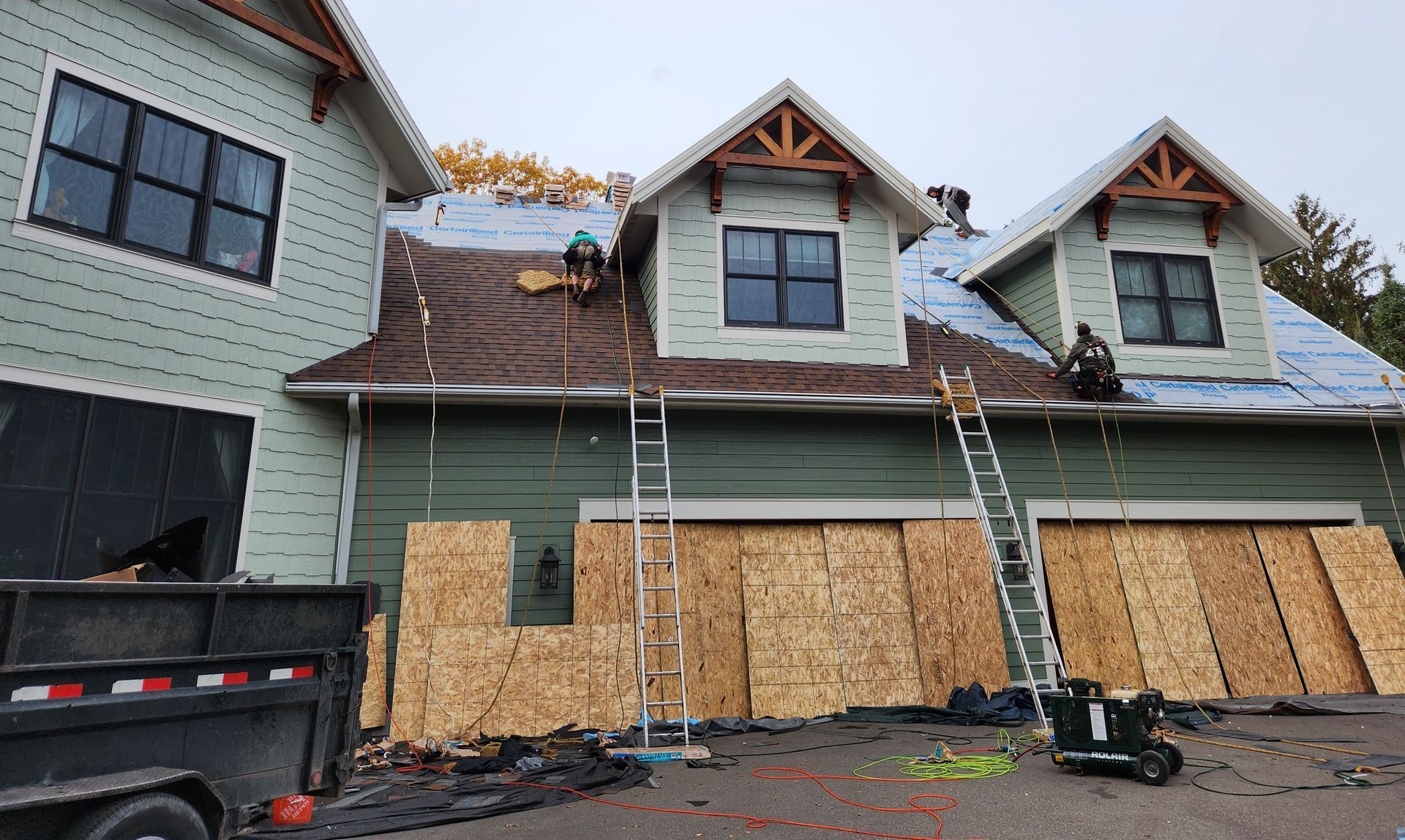 Workers installing a roof on a light green house with a garage. Boards cover the exterior walls.