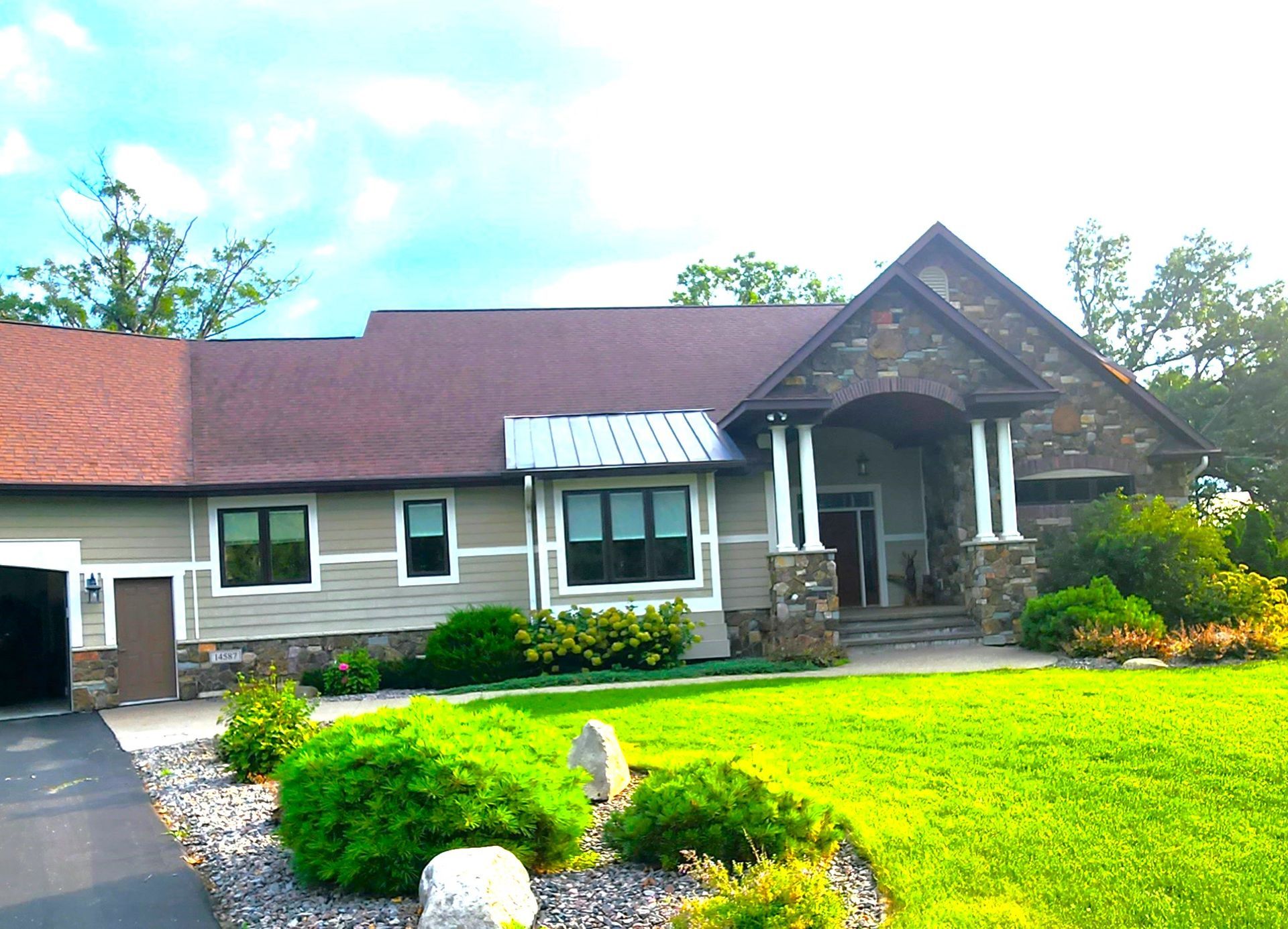 House with brown roof, stone facade, green lawn, and a driveway on a sunny day.