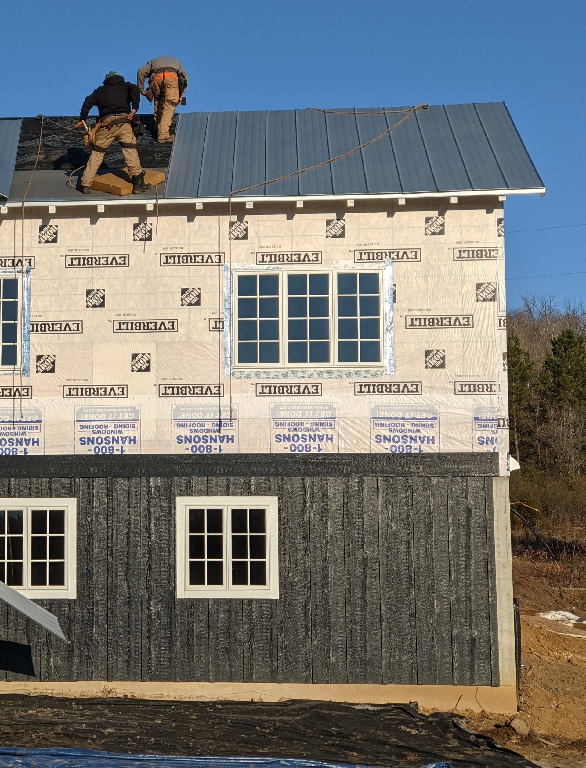 Construction workers install roofing on a partially built house with blue metal panels.