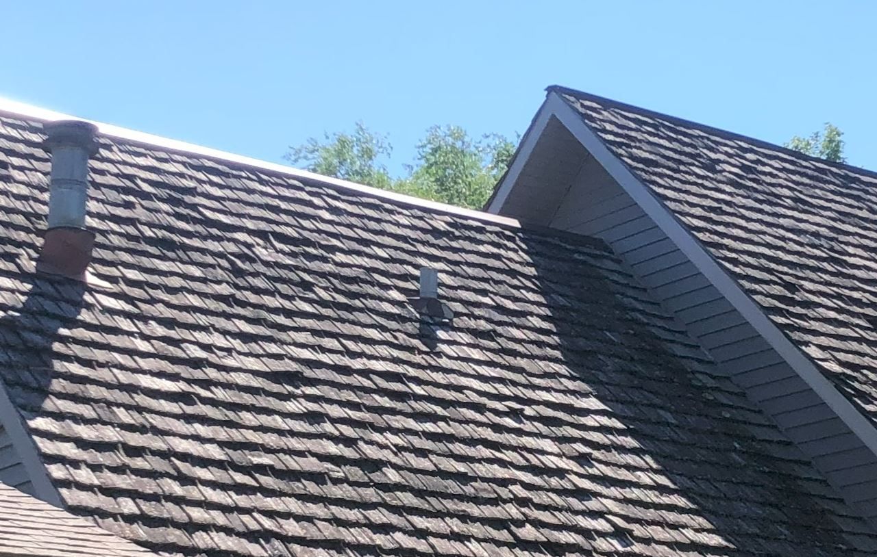 Cedar shake roof with chimney and vent pipe against a blue sky.