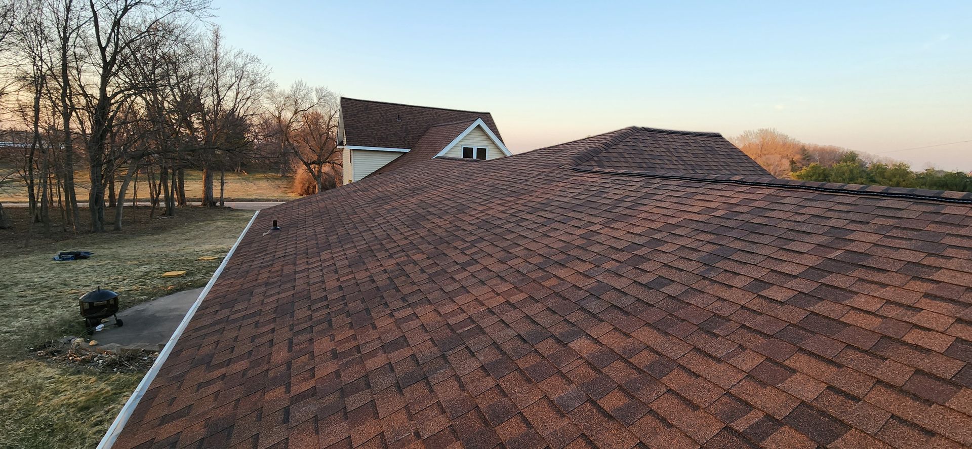 Brown shingle roof of a house with a view of the yard and trees. Blue sky in the background.