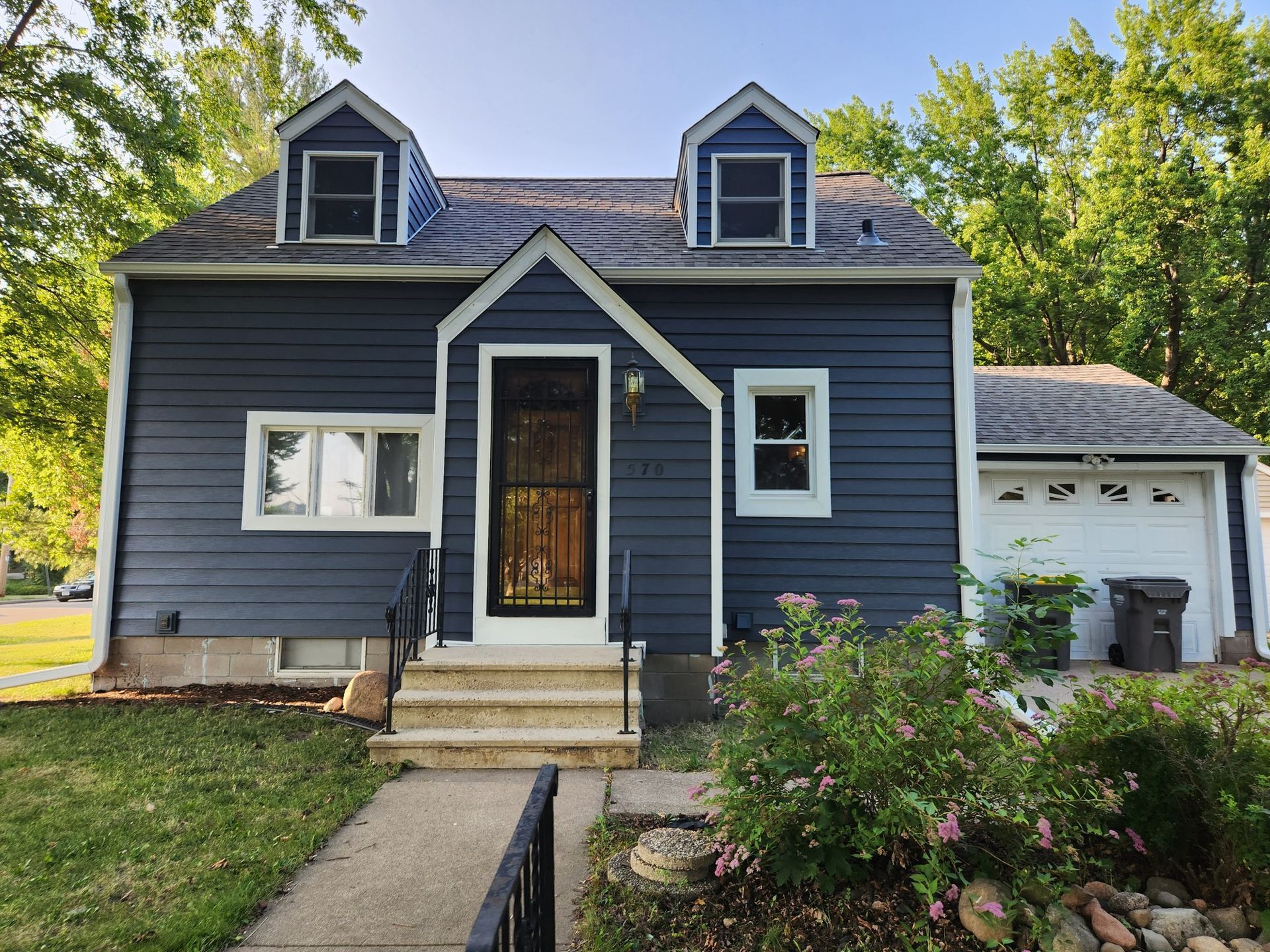 Blue house with white trim, two dormers, small front porch, attached garage, and green yard.