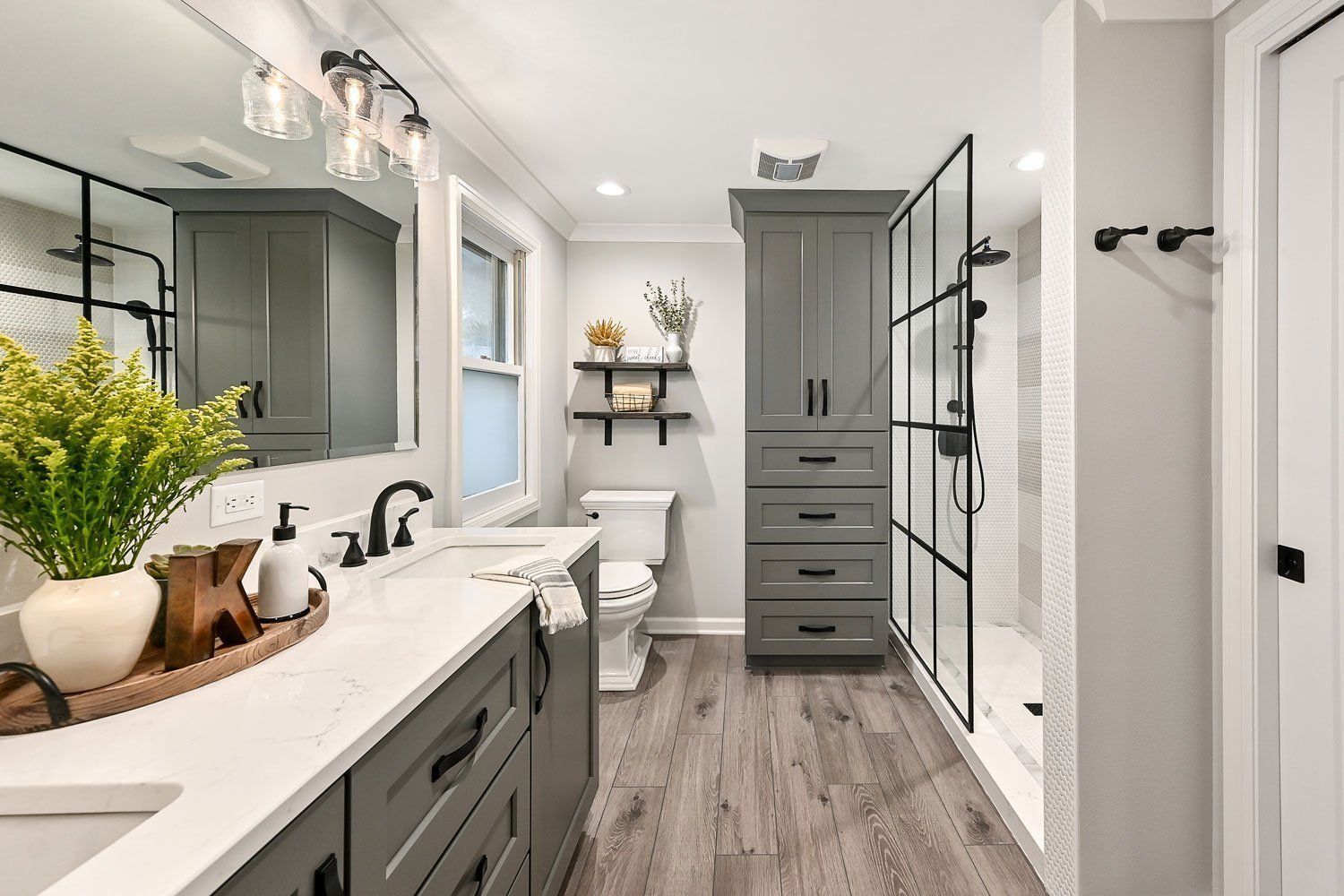 Modern bathroom with double vanity, gray cabinetry, glass shower, and white toilet in a bright, clean interior