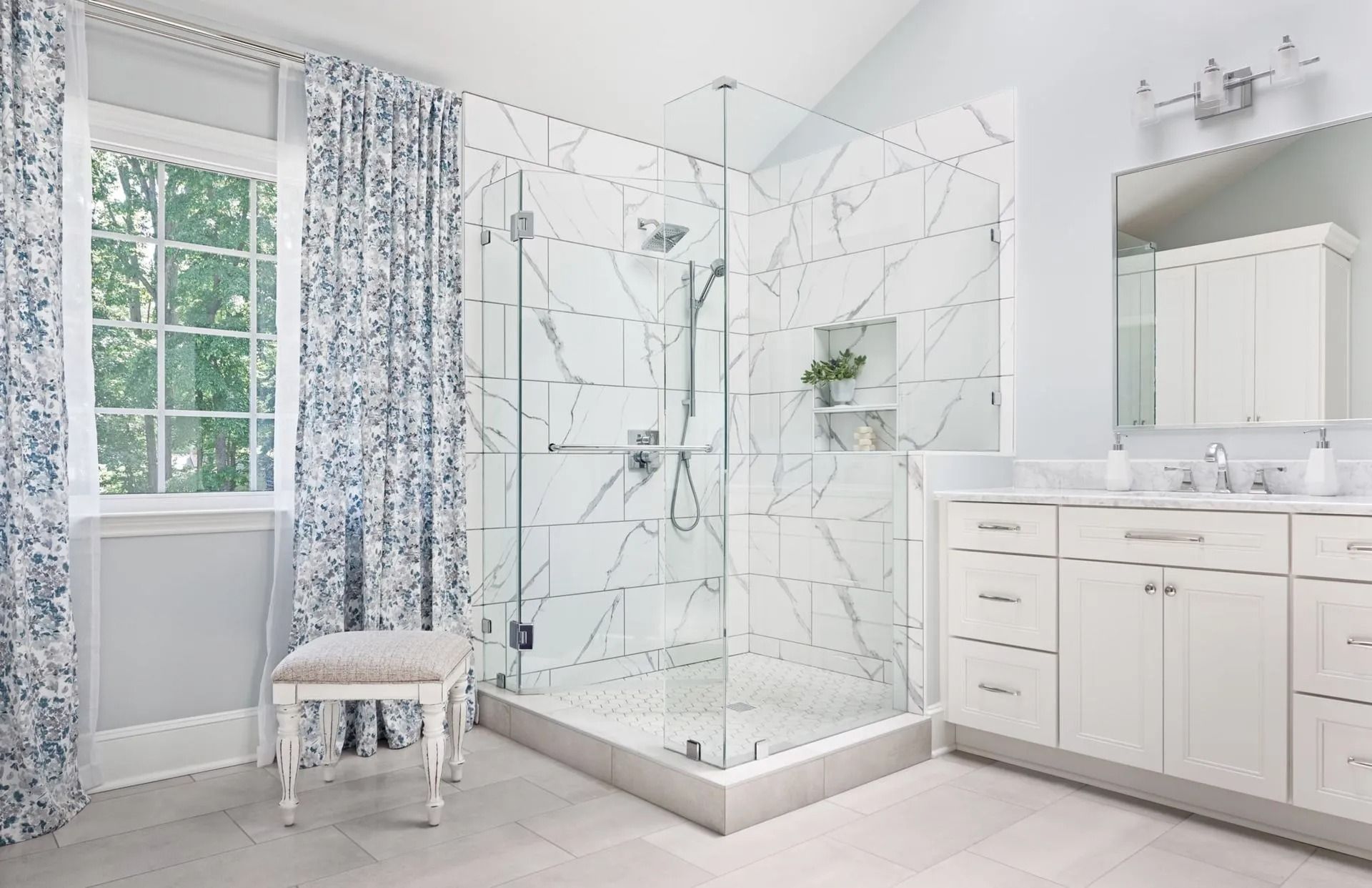 Bright white bathroom with glass shower, marble tile, vanity, window, and floral curtains