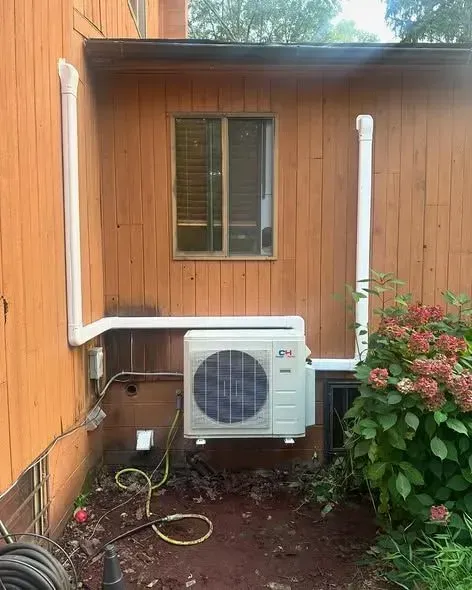 Outdoor wall-mounted air conditioner beside a wooden house with white piping and a small window.