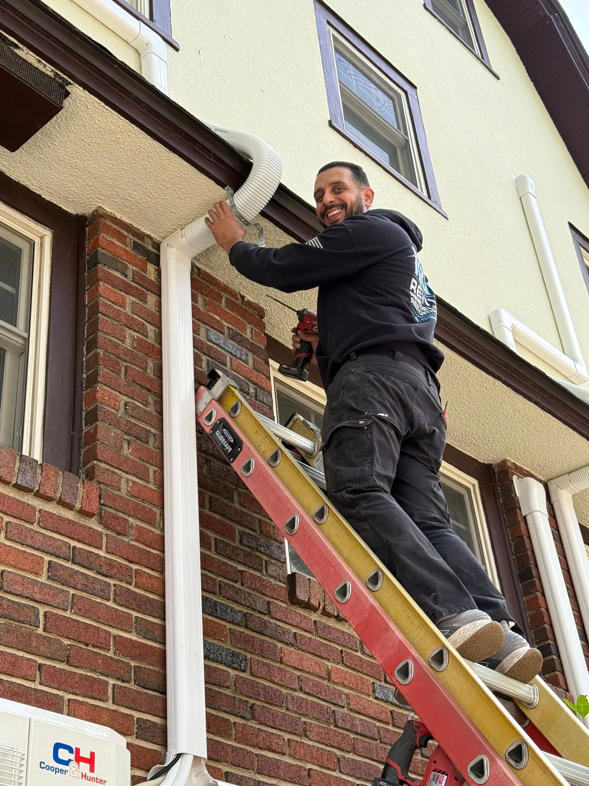 Technician on a ladder repairing a white gutter on a brick house exterior.