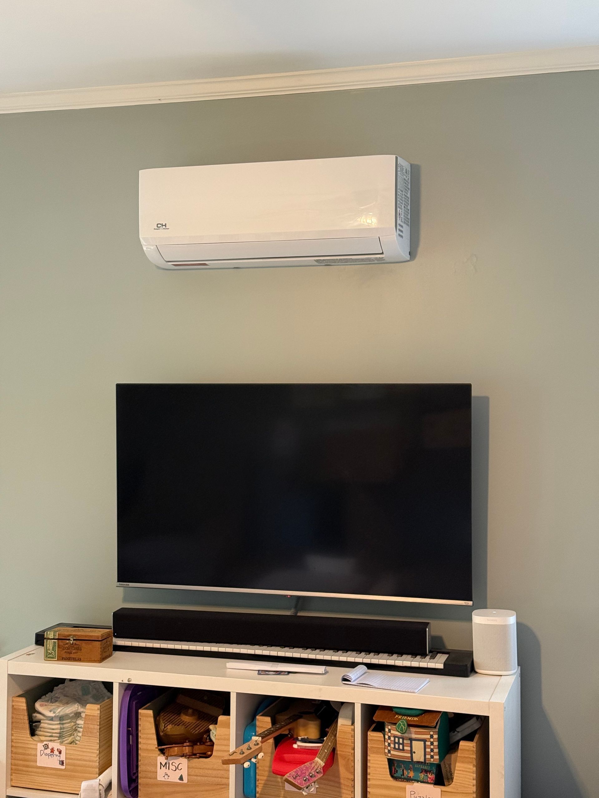 Wall-mounted white air conditioner above a black TV on a white stand with storage bins underneath
