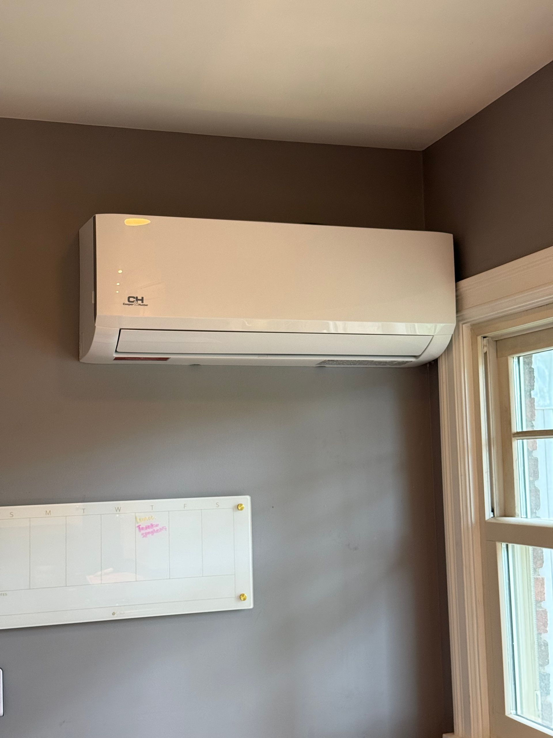 Wall-mounted white air conditioner above a whiteboard in a beige room beside a window.