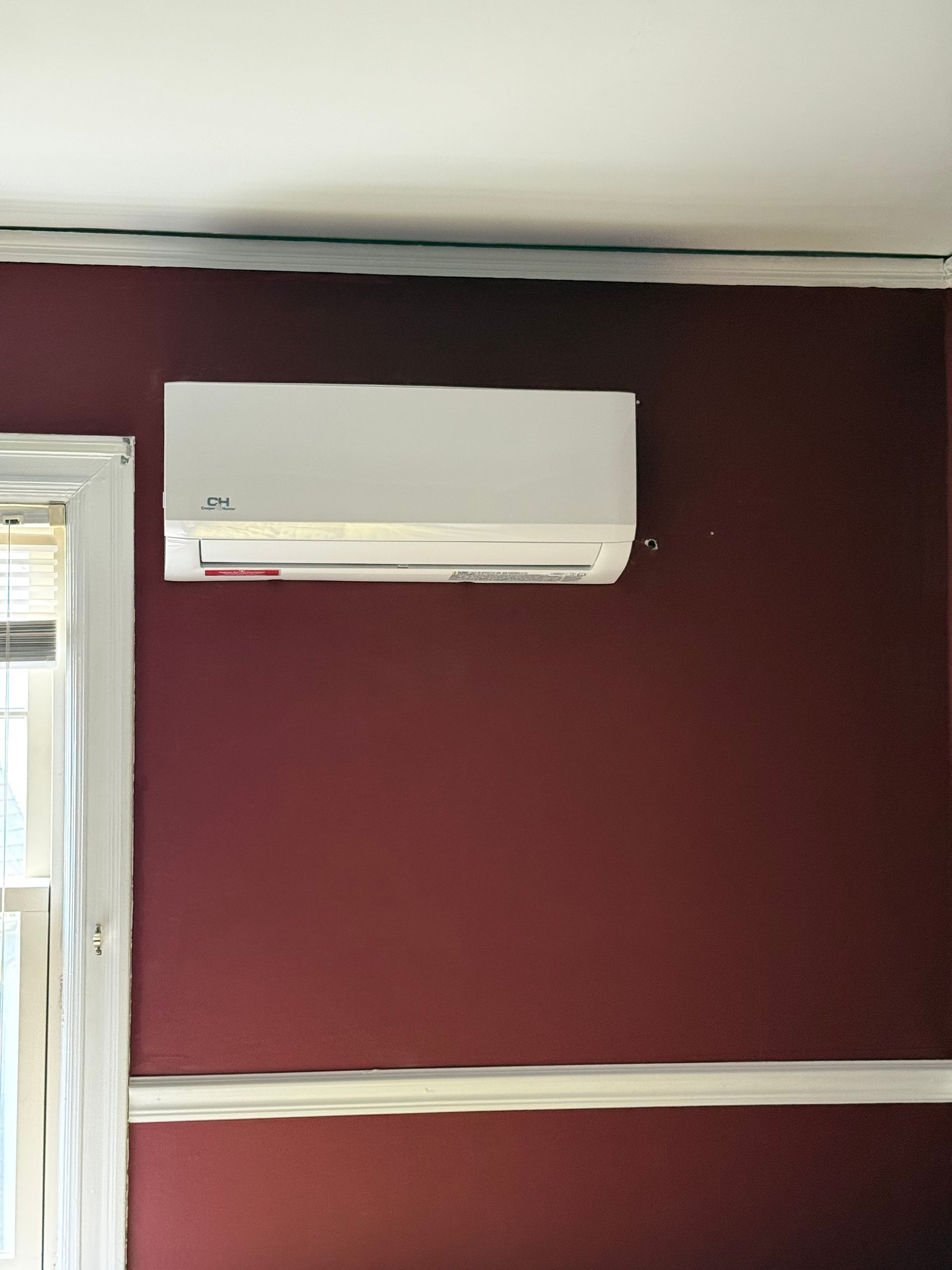 White wall-mounted air conditioner on a burgundy wall in a room with a window on the left.