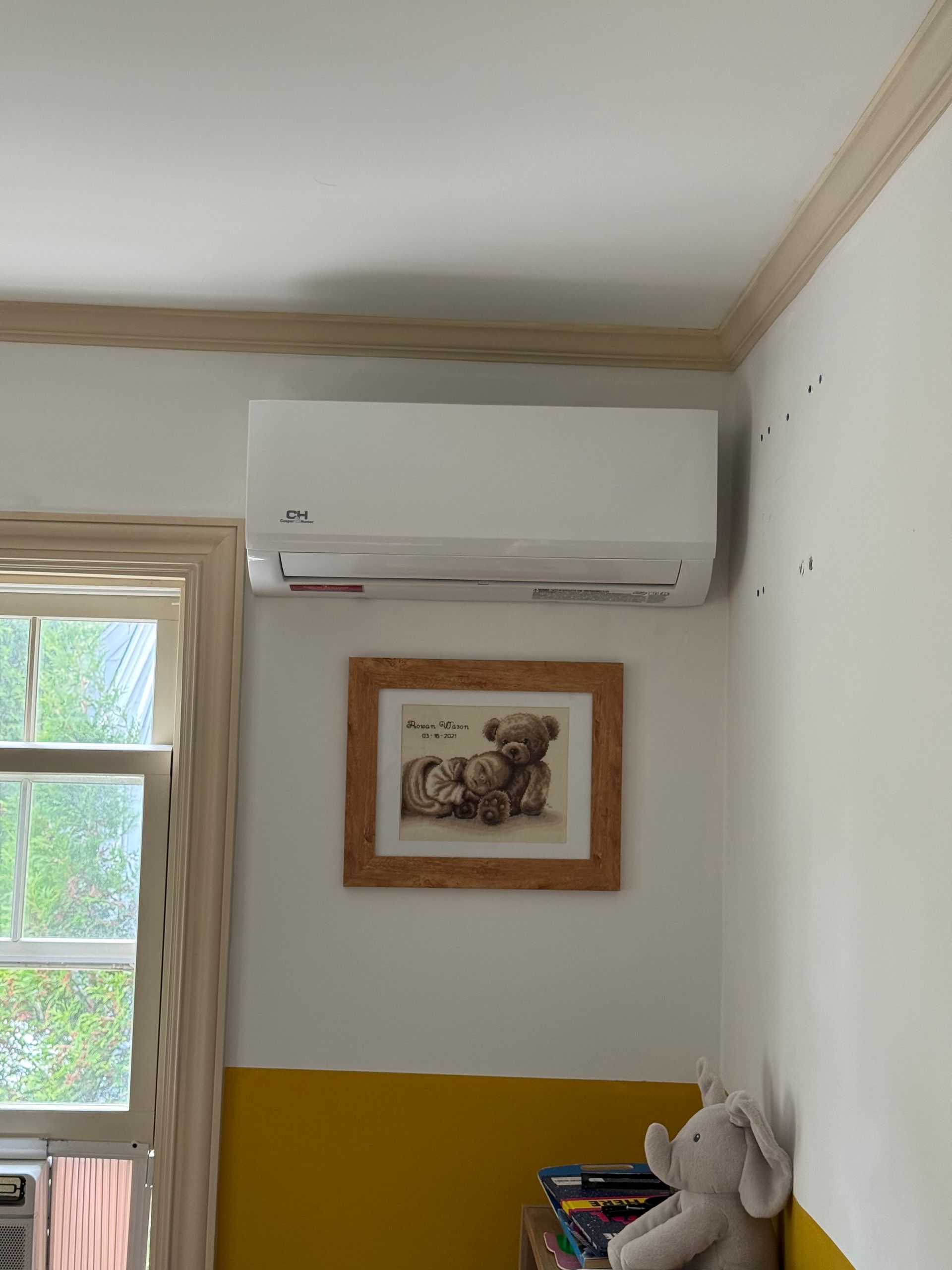 Wall-mounted air conditioner above a framed photo in a white room with a yellow lower wall and a window.