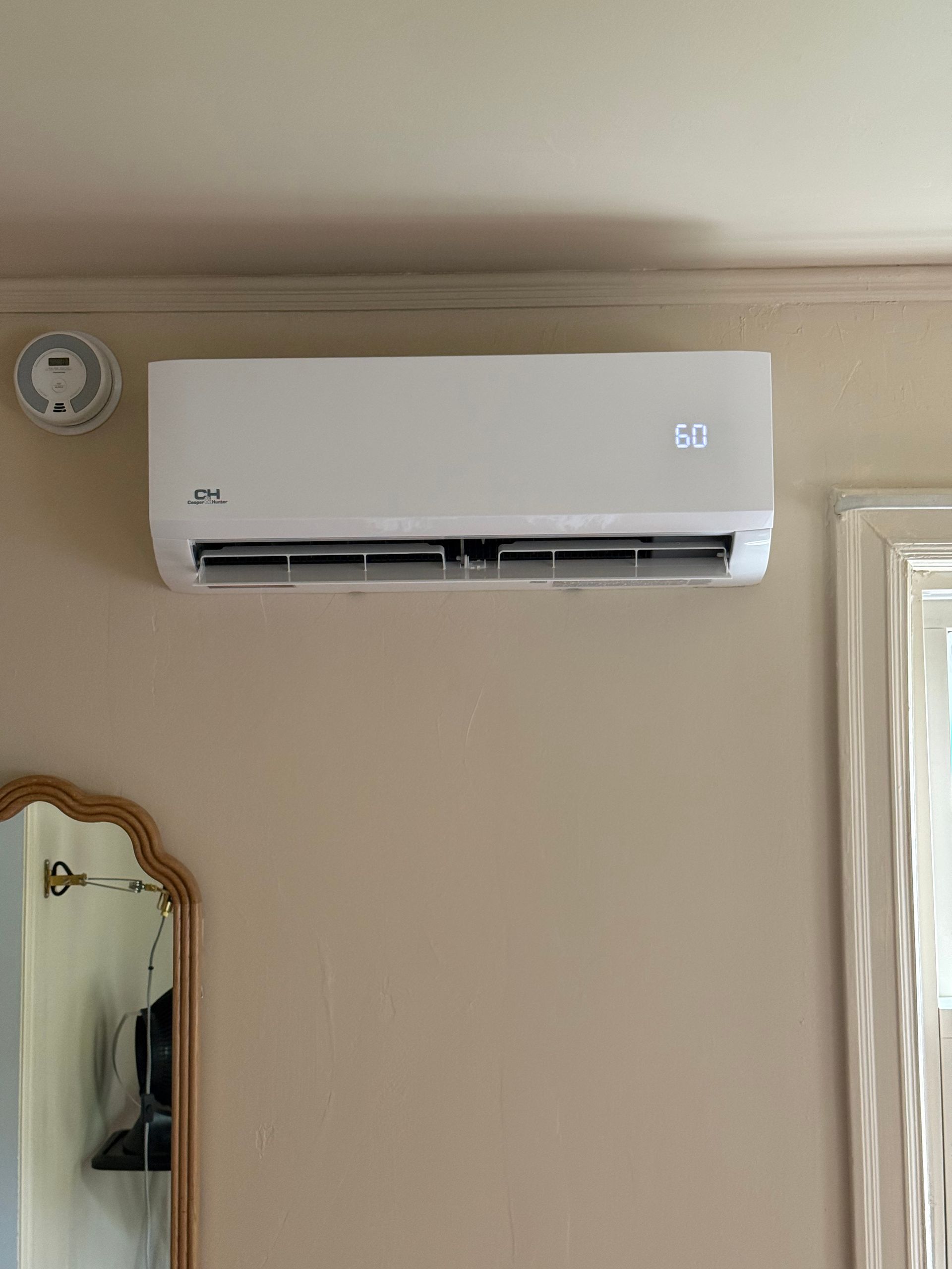 Wall-mounted white air conditioner above a beige wall, with a small clock and mirror nearby.